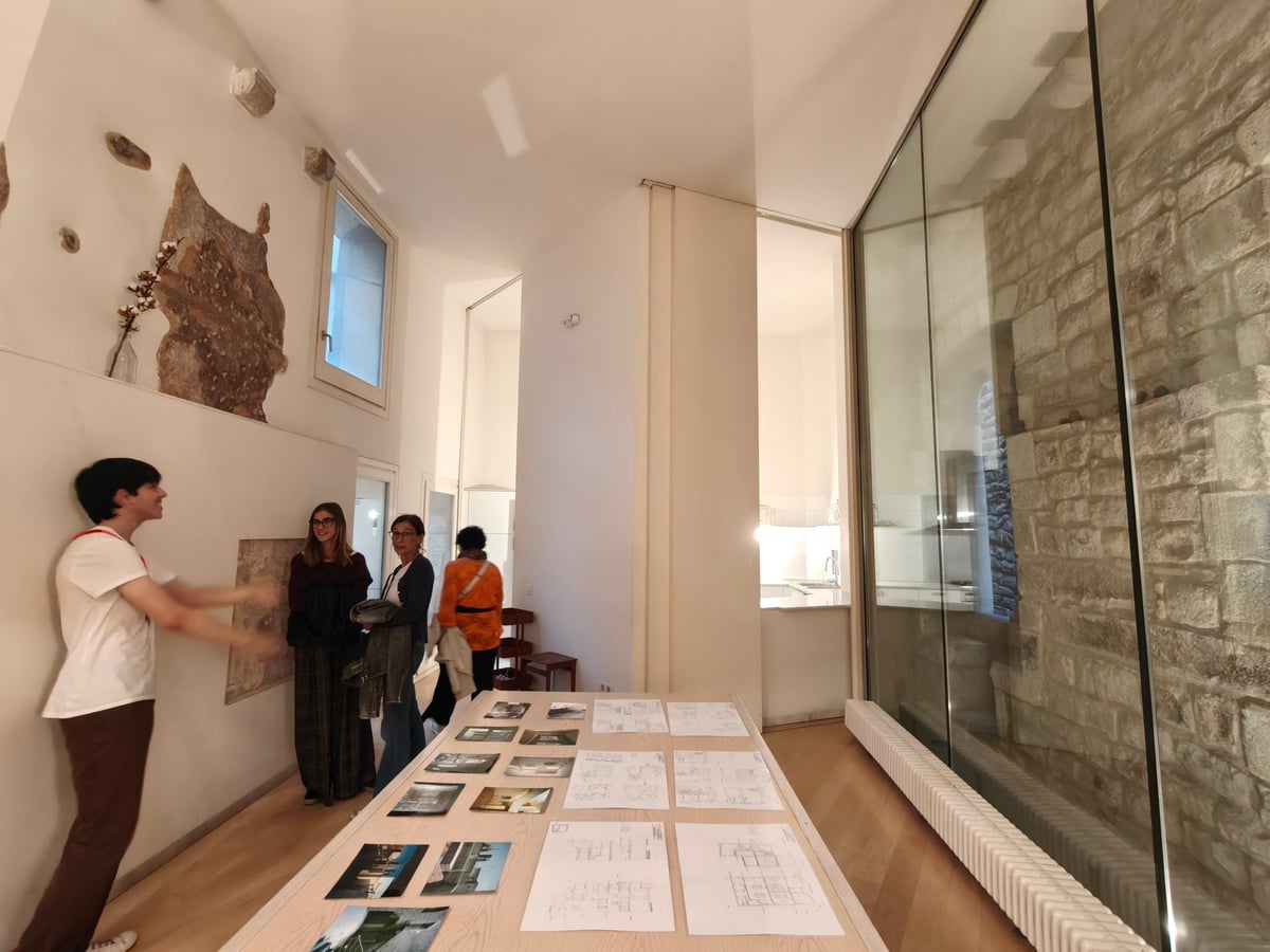 Sala de Casa Adret con un muro de piedra tras una cristalera, una mesa con fotografías y planos de la reforma y un grupo de visitantes atendiendo una explicación.