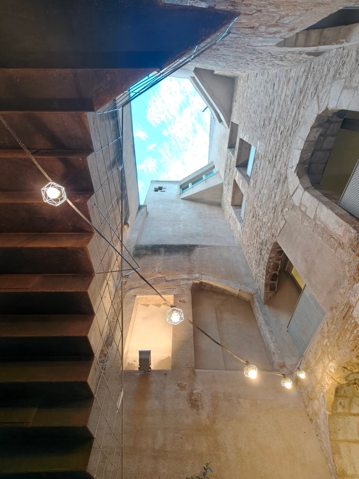 Patio de Casa Adret visto desde abajo, con muros de piedra, una estructura metálica oscura y hileras de bombillas hasta el cielo.