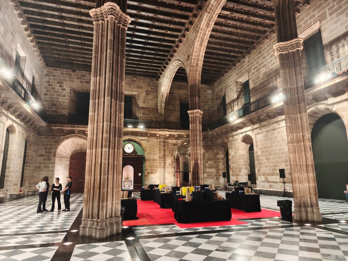 Sala gótica de la Llotja de Mar con grandes columnas de piedra, techo de vigas de madera, suelo de damero blanco y negro y mesas negras sobre una alfombra roja preparadas para la fiesta.