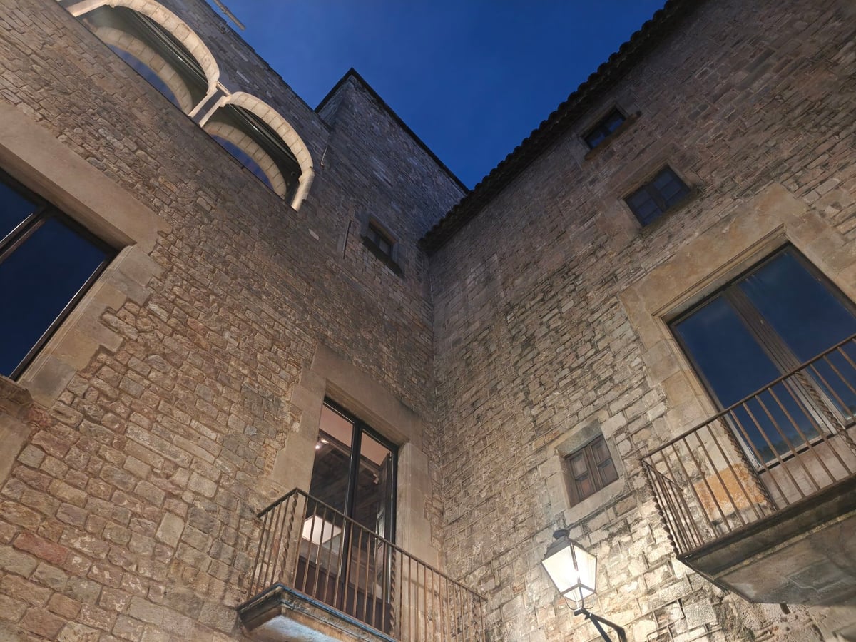 Muros de piedra de un patio gótico con balcones de hierro, ventanas y una farola, al anochecer.