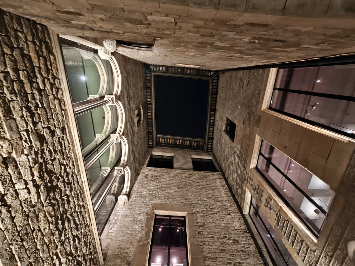 Patio de piedra visto desde abajo, con ventanales iluminados, galerías de arcos y un cuadrado de cielo oscuro arriba.