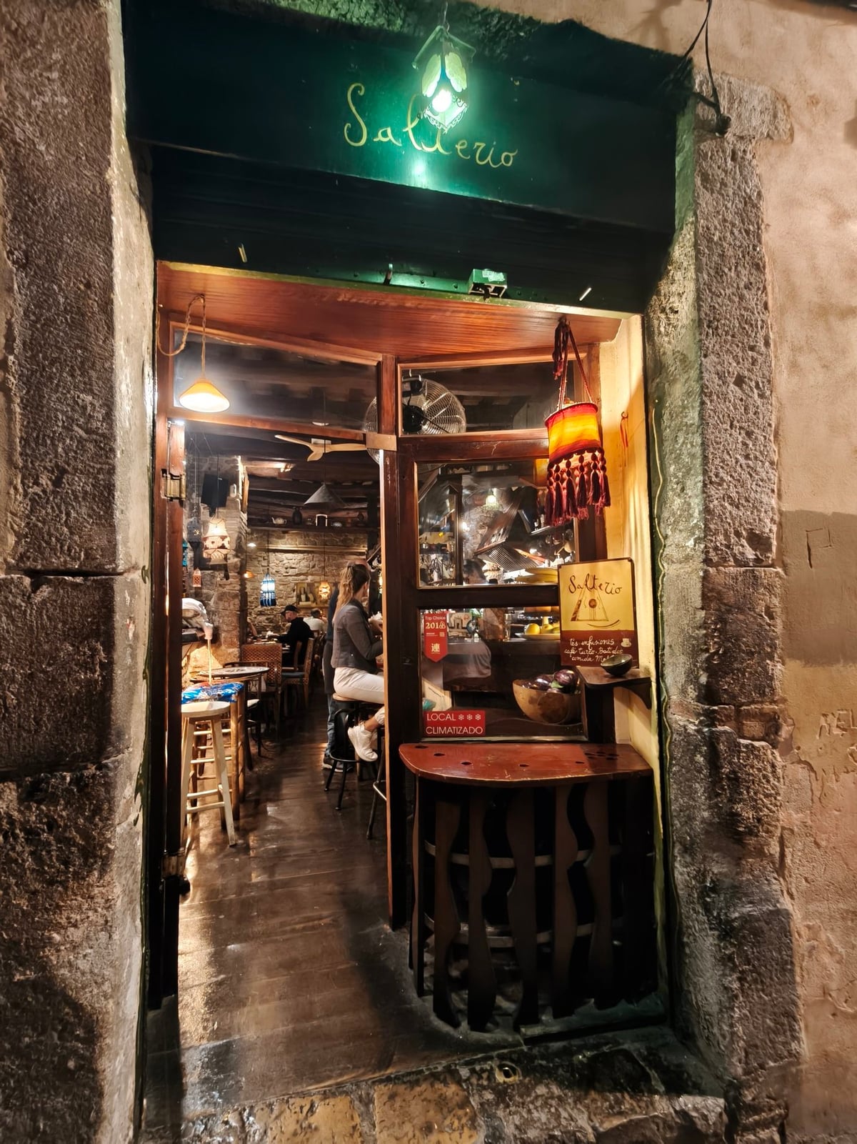 Entrada de Salterio de noche, con el rótulo pintado a mano sobre fondo verde, un farolillo encendido y, al fondo, el interior cálido con muros de piedra y mesas.