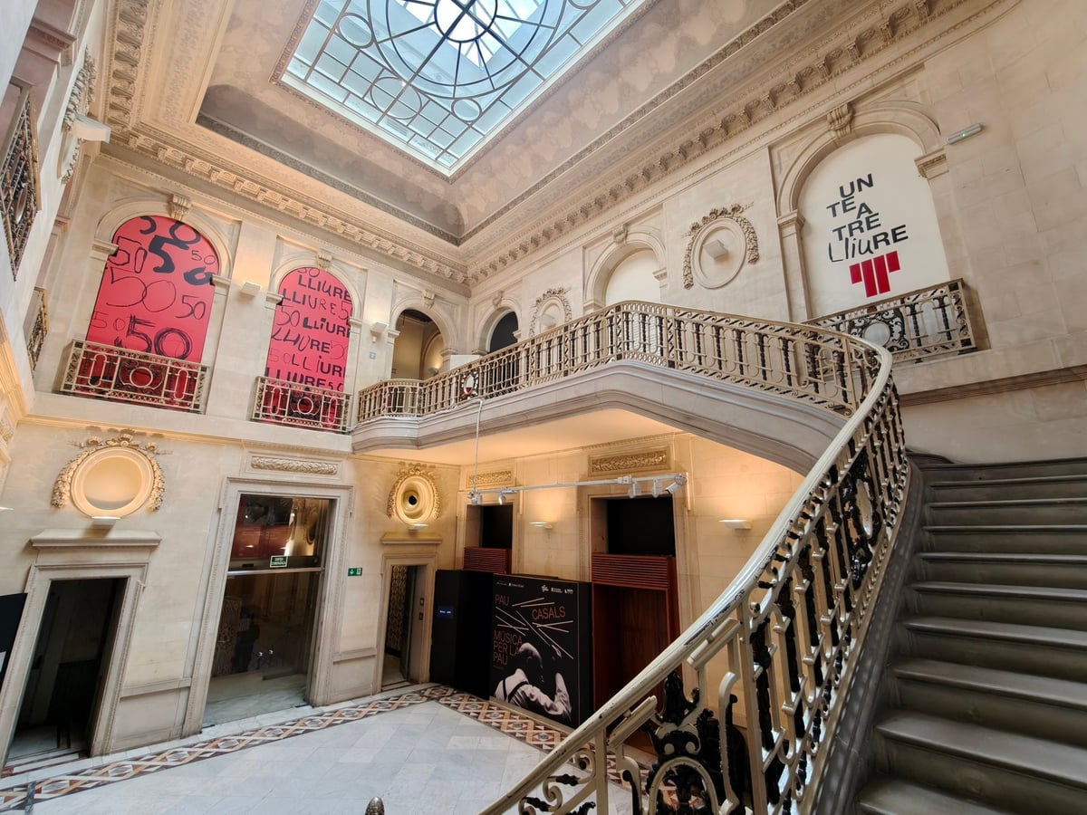 Escalera del Palau Robert, con barandilla de hierro, un lucernario de cristal y paños rosas con el título «Un teatre Lliure» colgados de los balcones.