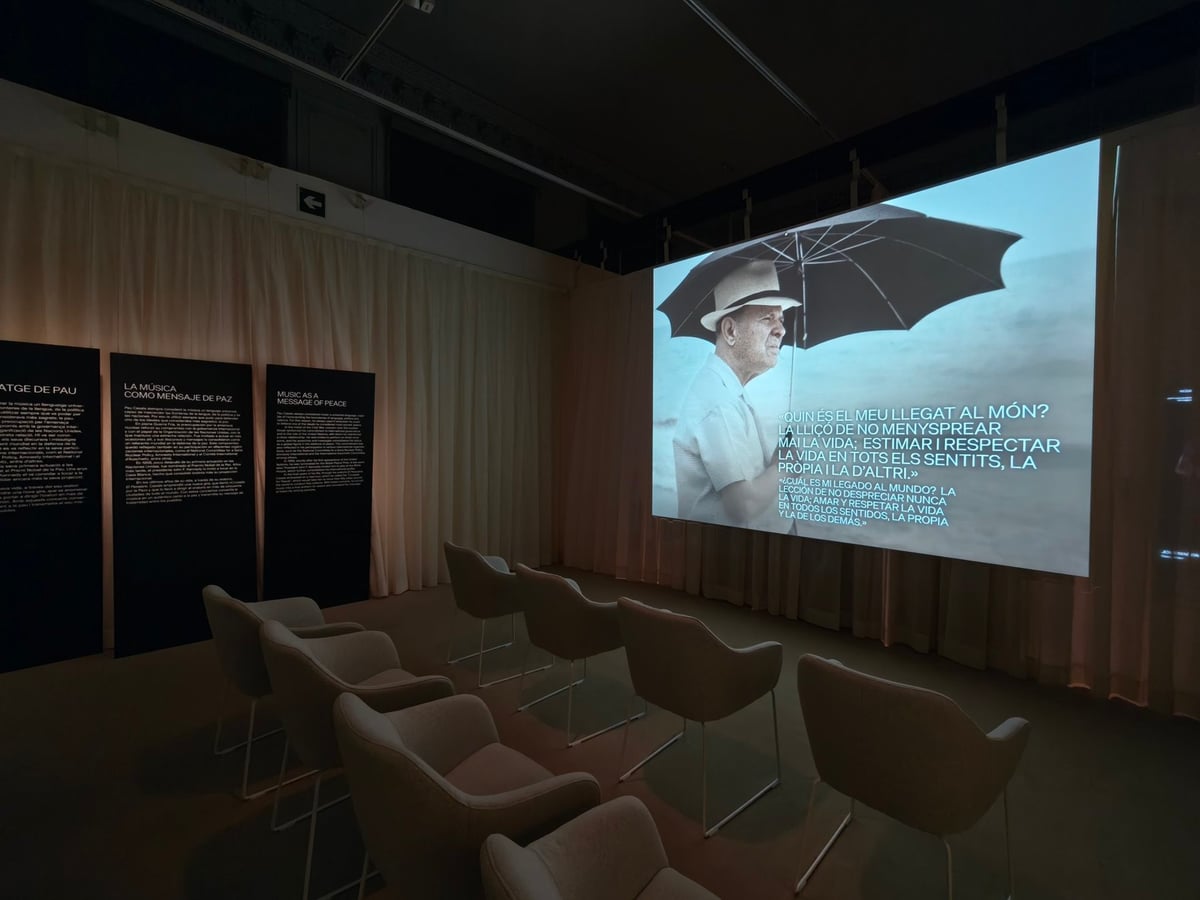 Sala oscura de la exposición sobre Pau Casals, con butacas frente a una pantalla donde se proyecta a un hombre mayor con sombrero bajo una sombrilla, y paneles de texto en catalán.