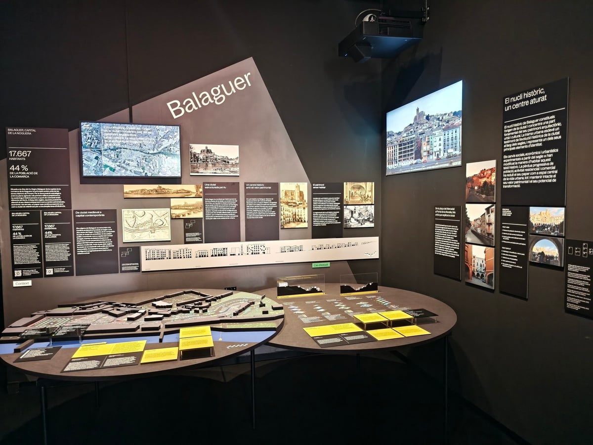 Mesa con maquetas y planos del casco histórico de Balaguer ante un panel con fotografías, datos y una pantalla, en la exposición «Ciutats en trànsit».