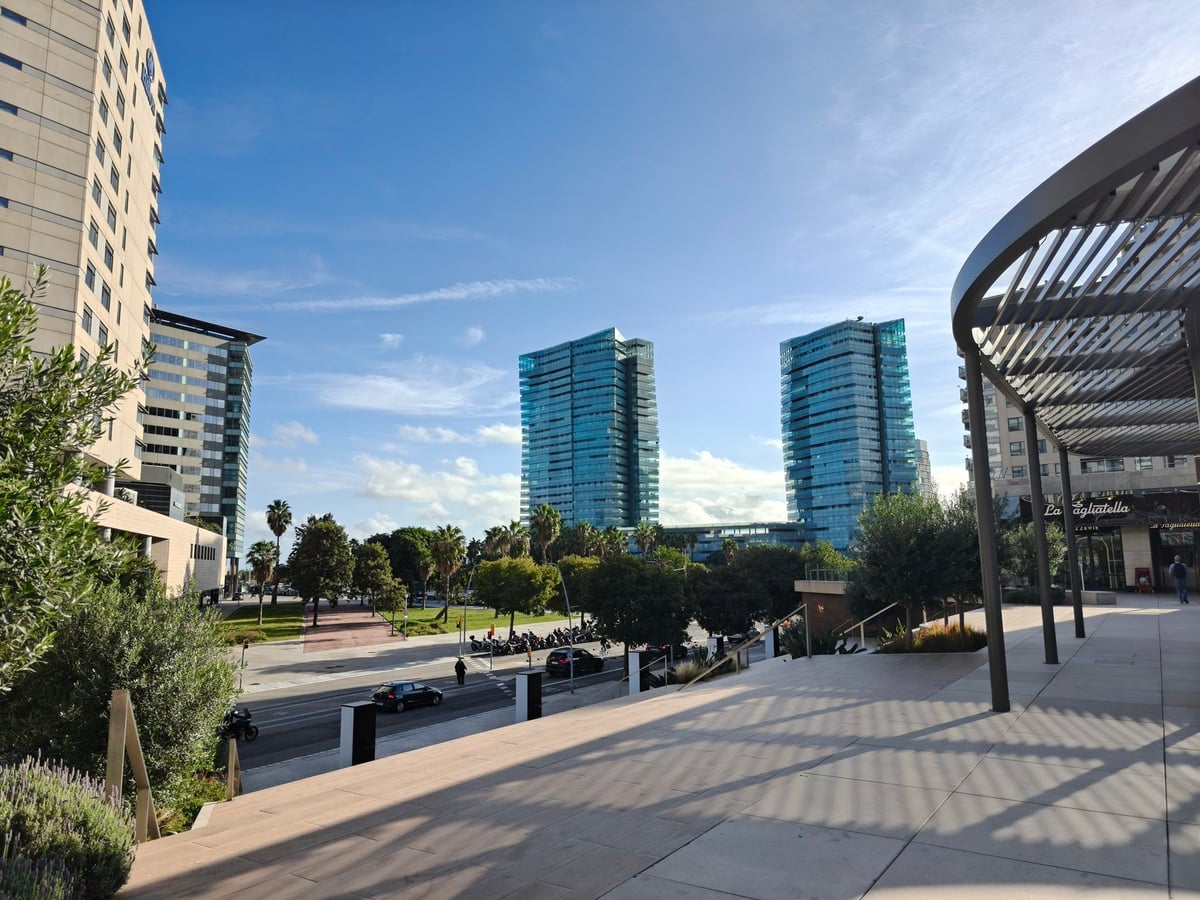 Dos torres de cristal azul en Diagonal Mar vistas desde una explanada con una pérgola metálica, bajo un cielo con nubes.