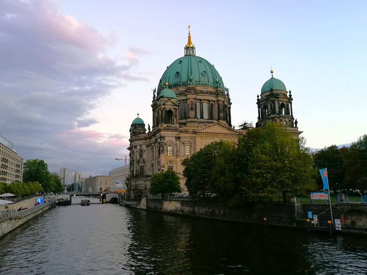La catedral de Berlín al atardecer vista desde el río Spree, con su gran cúpula de cobre verde y la linterna dorada, el agua oscura en primer plano y un barco turístico pasando bajo un puente al fondo.