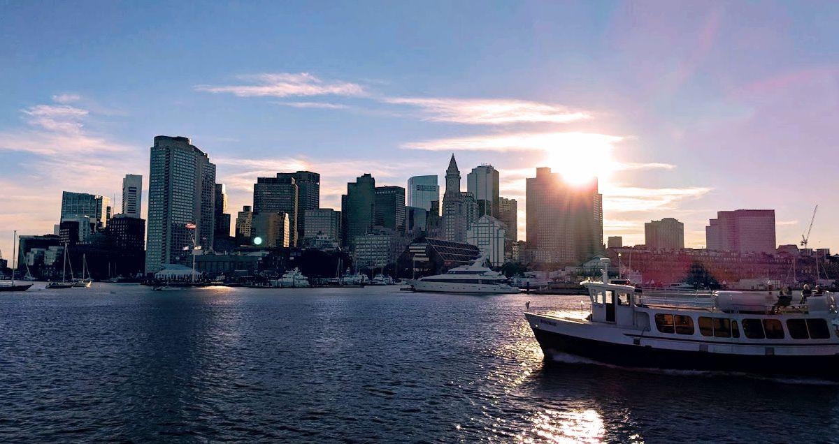 El skyline de Boston al atardecer visto desde el agua, con los rascacielos recortados a contraluz, el sol colándose entre dos torres y un pequeño ferry blanco cruzando el puerto en primer plano.