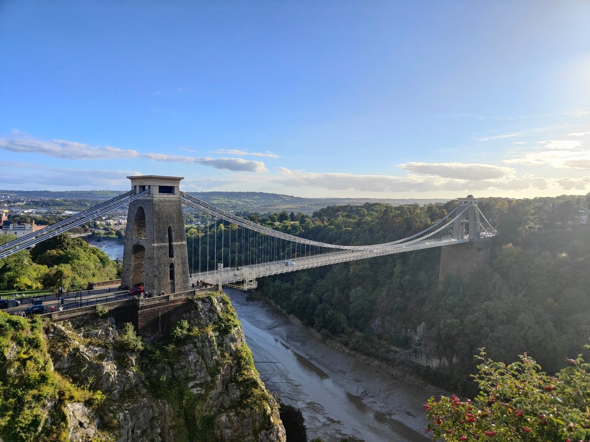 El puente colgante de Clifton tendido sobre la garganta del río Avon, con la torre de piedra en primer plano sobre el acantilado, el cauce embarrado abajo y la campiña inglesa perdiéndose en el horizonte.