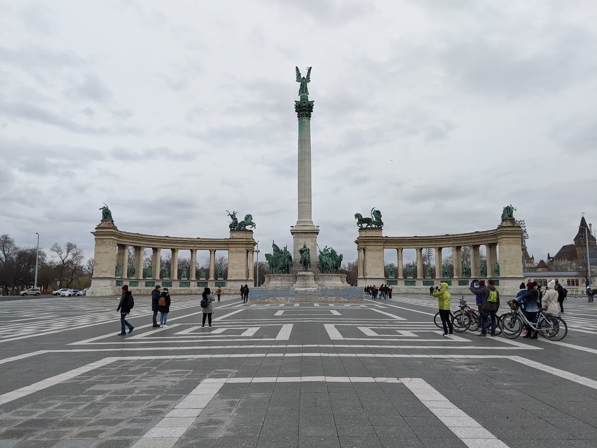 La plaza de los Héroes de Budapest un día nublado, con la columna coronada por el arcángel Gabriel en el centro, las dos columnatas semicirculares llenas de estatuas de bronce a los lados y unos pocos visitantes cruzando la explanada.