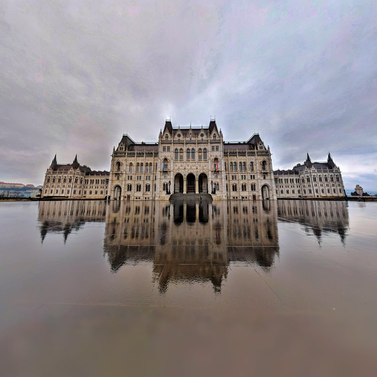 La fachada neogótica del Parlamento de Budapest vista de frente y reflejada casi entera en el suelo mojado de la plaza, bajo un cielo encapotado de nubes malvas