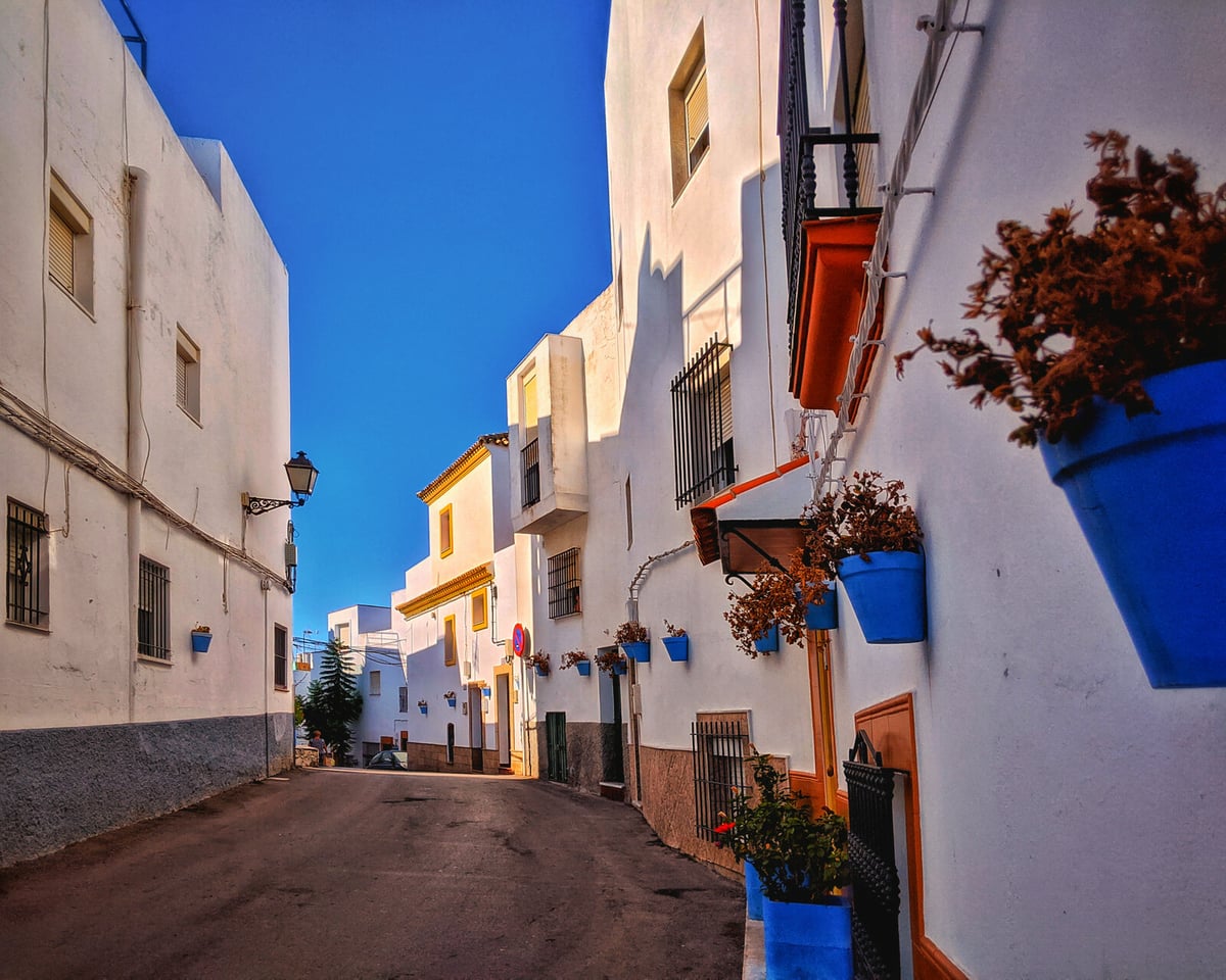 Calle en cuesta de un pueblo andaluz, con las casas encaladas de blanco recortadas contra un cielo azul intenso, macetas azules colgadas de las fachadas y rejas de forja en las ventanas.