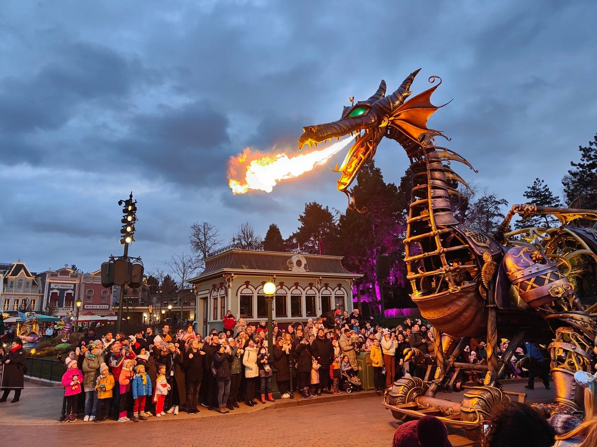 El dragón mecánico de la cabalgata nocturna de Disneyland París escupiendo una llamarada real por la boca, con el público agolpado tras las vallas grabándolo con los móviles al anochecer.