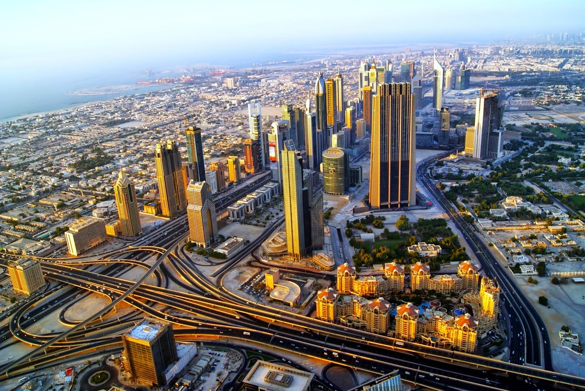 Dubái a vista de pájaro desde lo alto de un rascacielos al atardecer, con las torres de la Sheikh Zayed Road formando un corredor iluminado, los nudos de autopistas serpenteando abajo y el golfo al fondo.