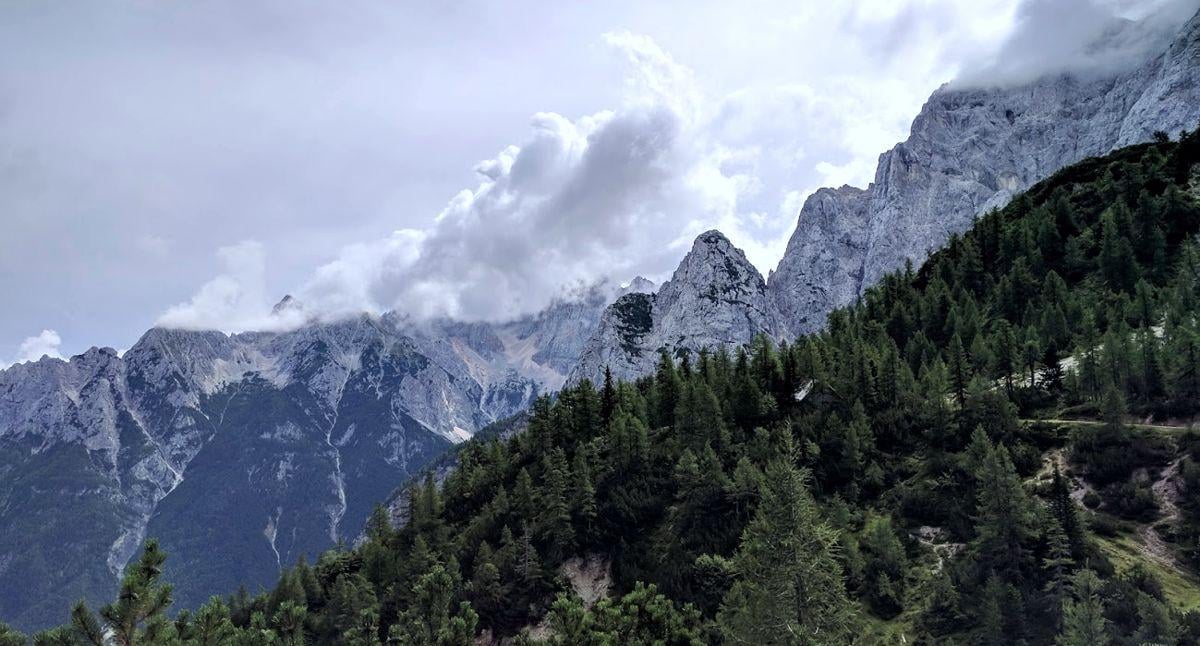 Una de las curvas adoquinadas del paso Vršič ascendiendo entre bosque de coníferas, con las cumbres de los Alpes Julianos al fondo.