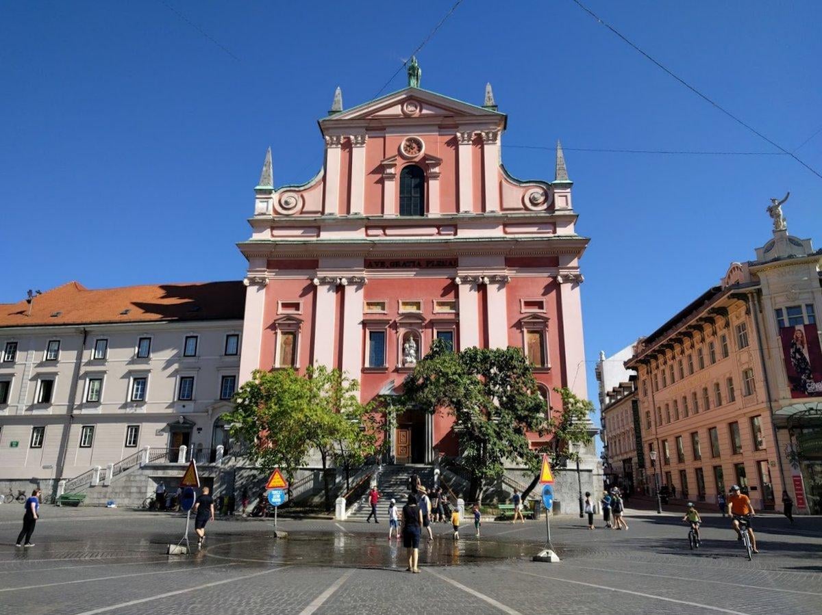 El Puente Triple de Ljubljana sobre el río Ljubljanica, con la fachada roja de la Iglesia Franciscana al fondo y las terrazas de la ribera a ambos lados.