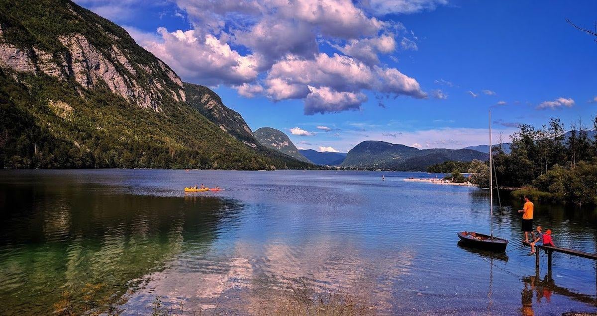 El lago de Bohinj en calma, con las montañas boscosas de los Alpes Julianos reflejadas en el agua, una canoa amarilla en el centro y un pequeño velero amarrado junto a un embarcadero de madera.
