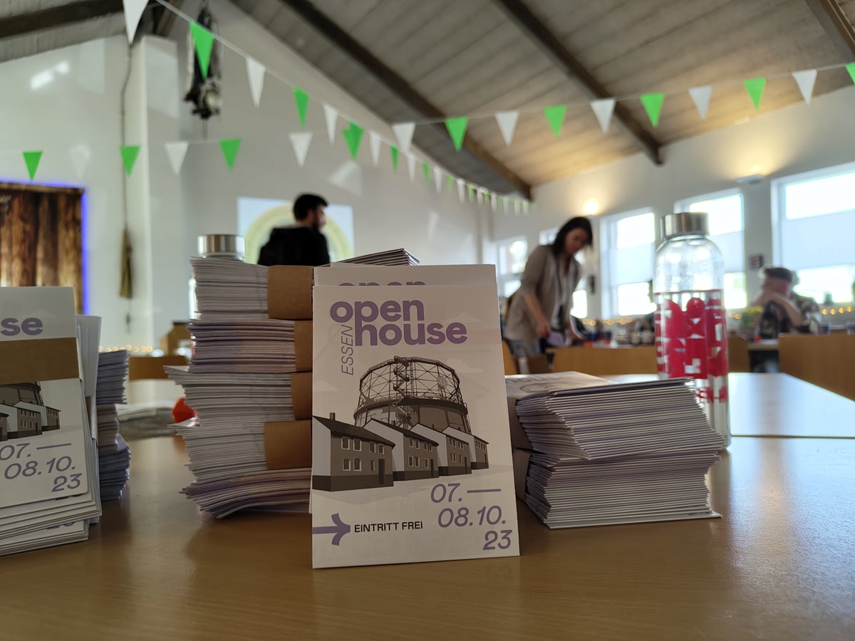 Montones de programas del Open House de Essen apilados sobre una mesa, con uno de pie en primer plano que muestra un gasómetro dibujado sobre una hilera de casas obreras, y voluntarios preparando el puesto al fondo bajo guirnaldas de banderines verdes.