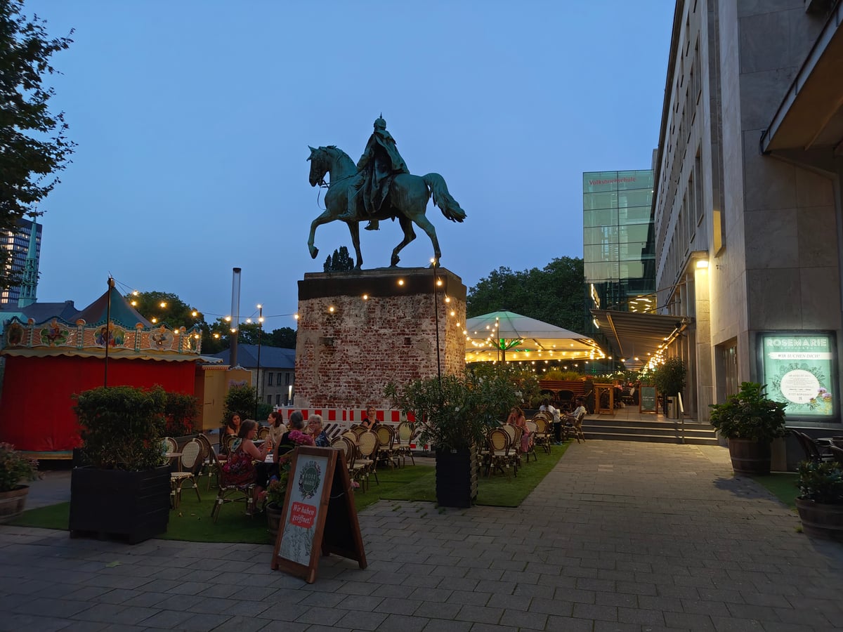 Una estatua ecuestre de bronce sobre un pedestal de ladrillo al anochecer, rodeada de guirnaldas de bombillas, con las sillas de mimbre de una terraza a sus pies y un tiovivo de feria iluminado a la izquierda.
