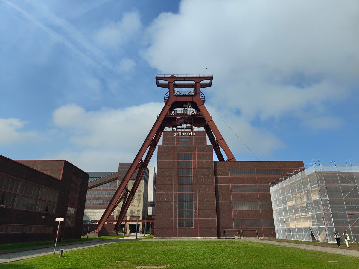 El castillete rojo de la mina Zollverein de Essen, una torre de acero en forma de A con las dos poleas de extracción en lo alto, sobre los edificios de ladrillo del complejo minero bajo un cielo azul.
