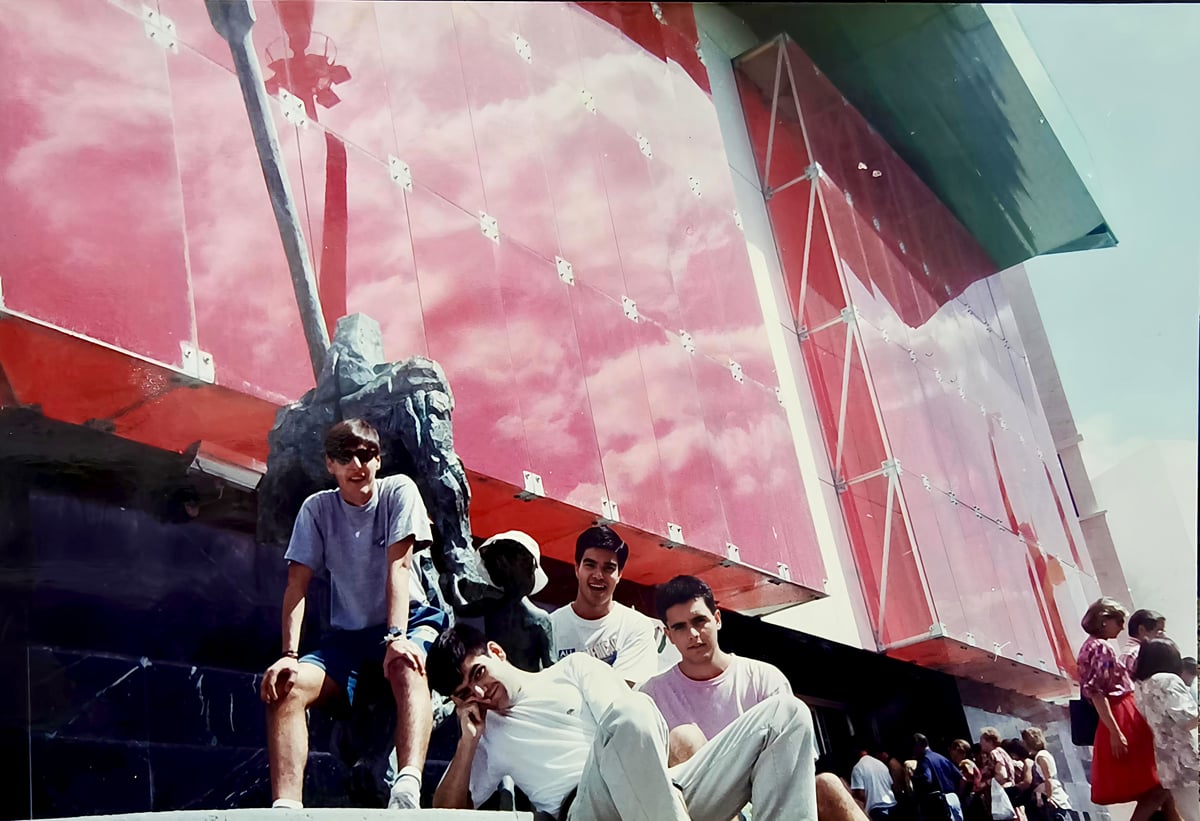 Cuatro chavales posan sentados en unas escaleras junto a una escultura de bronce, delante de la fachada de vidrio rojo de un pabellón de la Expo 92, en una foto de carrete de los años noventa.