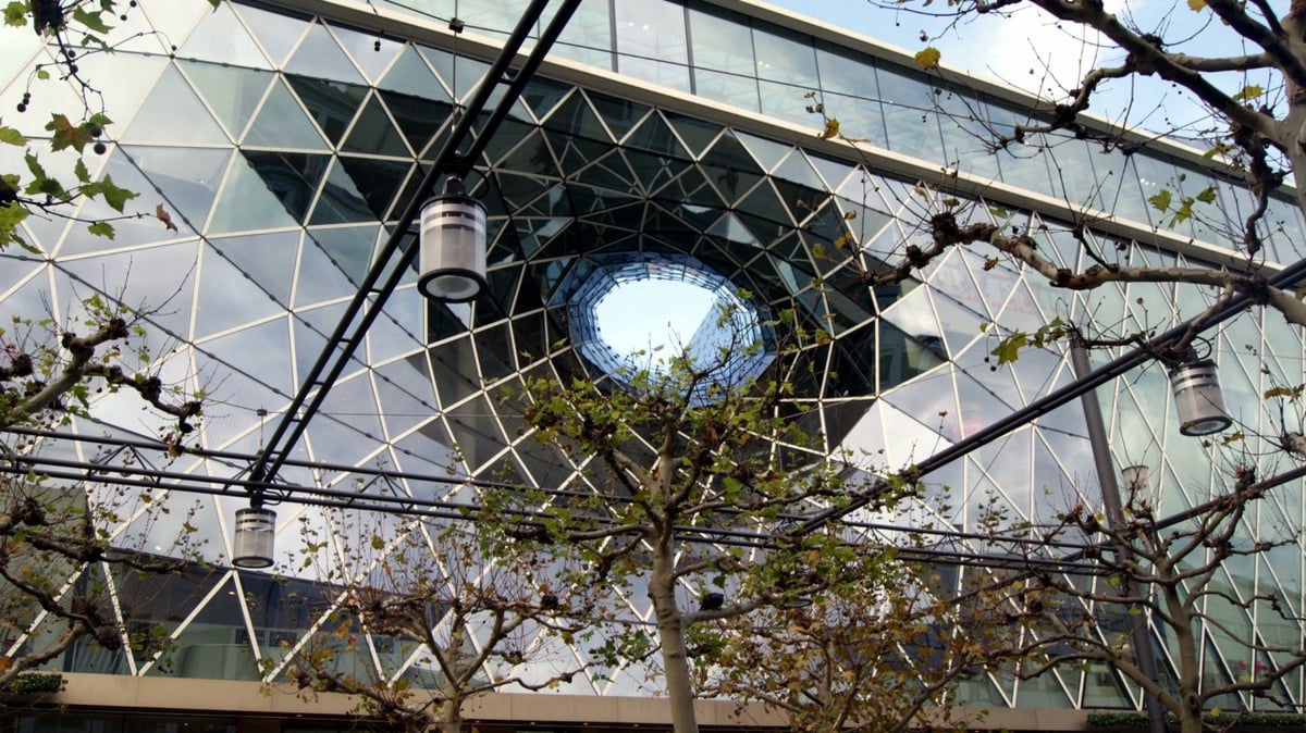 La fachada de vidrio triangulado del centro comercial MyZeil de Fráncfort, con el gran embudo acristalado que se hunde en su interior, vista entre las ramas peladas de los árboles de la calle.