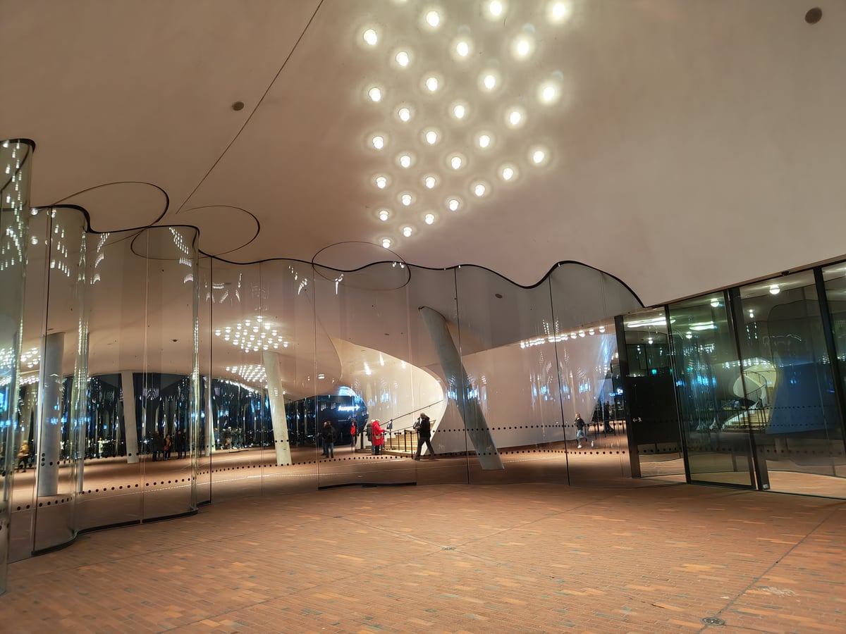 La plaza pública de la Elbphilharmonie de Hamburgo, con su techo blanco salpicado de luminarias circulares y un muro ondulado de vidrio curvo que separa el mirador del vestíbulo iluminado.