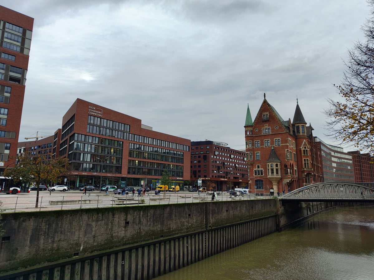 La HafenCity de Hamburgo desde el muelle, con un edificio moderno de ladrillo y cristal a la izquierda, una casa neogótica de torres puntiagudas a la derecha y un puente de hierro cruzando el canal.