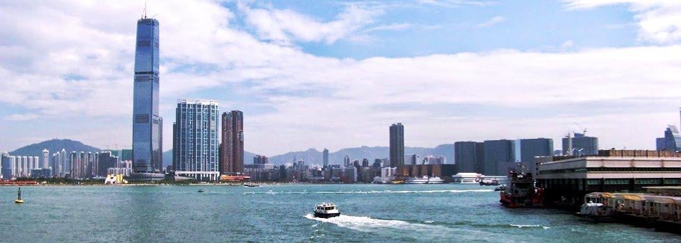 El puerto de Hong Kong desde el paseo marítimo, con la torre acristalada del International Commerce Centre destacando sobre los edificios de Kowloon, una lancha cruzando el agua y un embarcadero con barcos amarrados a la derecha.