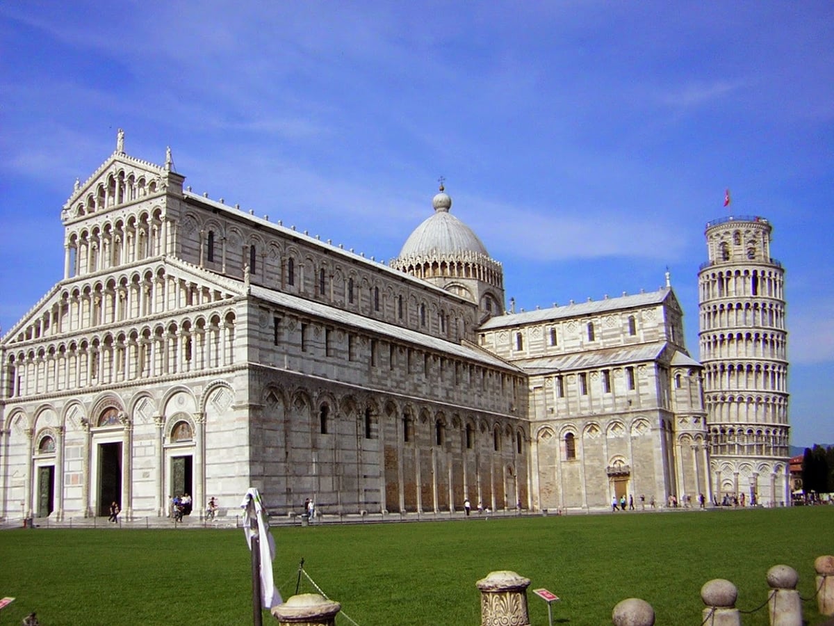La catedral de Pisa en mármol blanco y gris a franjas, con su cúpula ovalada, vista desde el césped de la plaza de los Milagros, con la torre inclinada asomando a la derecha bajo un cielo azul.