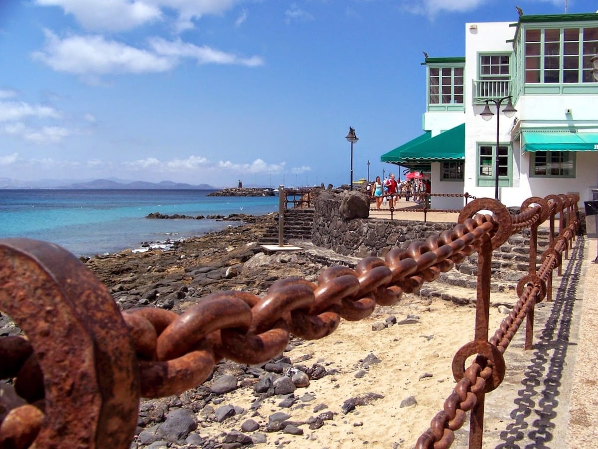 Paseo marítimo de un pueblo costero de Lanzarote, con una cadena de hierro oxidada haciendo de barandilla en primer plano, la orilla de roca volcánica y arena, el agua turquesa y una casa blanca de toldos verdes.