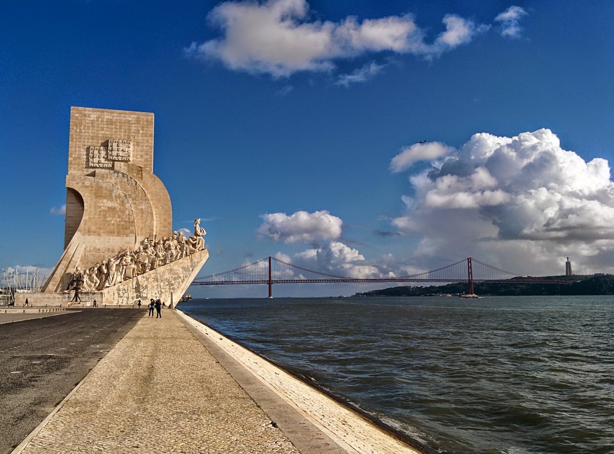 El Monumento a los Descubrimientos de Lisboa visto desde el muelle, con su proa de piedra cargada de figuras mirando al Tajo, el puente 25 de Abril al fondo y grandes nubes blancas sobre el río.