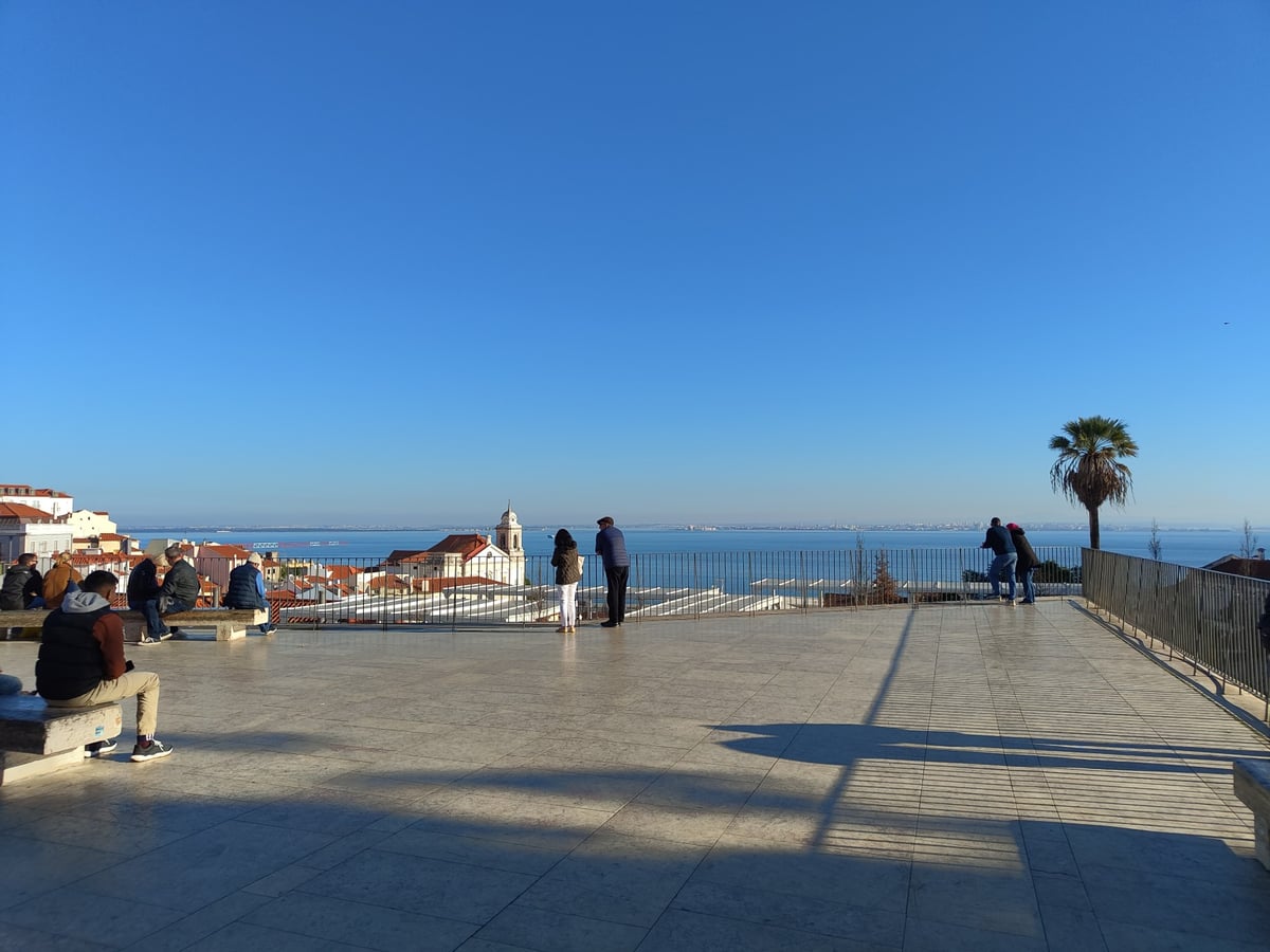 Vista desde un mirador de Alfama con los tejados del barrio bajando hacia el Tajo, la cúpula blanca del Panteón a la izquierda y el río abriéndose al fondo.
