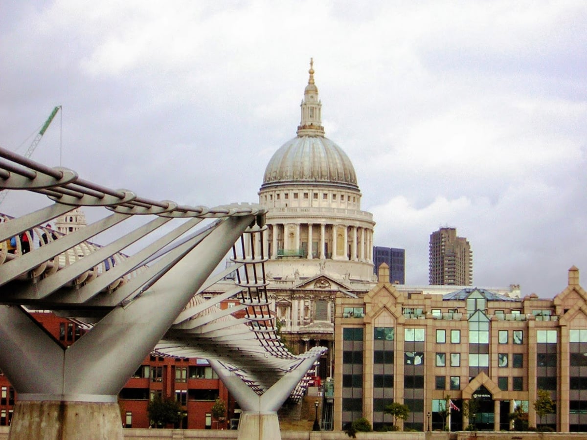 La cúpula de la catedral de San Pablo de Londres bajo un cielo gris, vista desde la orilla sur del Támesis, con la estructura de acero del Millennium Bridge cruzando en primer plano.