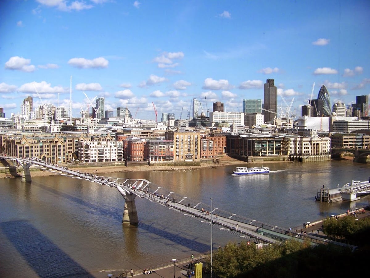 El Támesis desde lo alto, con el Millennium Bridge cruzando en diagonal lleno de gente, los almacenes victorianos de la orilla norte y los rascacielos de la City al fondo bajo un cielo azul con nubes.