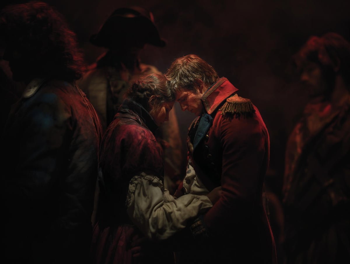Dos personajes de Los Miserables juntan las frentes en un abrazo bajo una luz rojiza, ella con vestido raído y él con casaca militar roja y charreteras doradas, rodeados de figuras en penumbra.