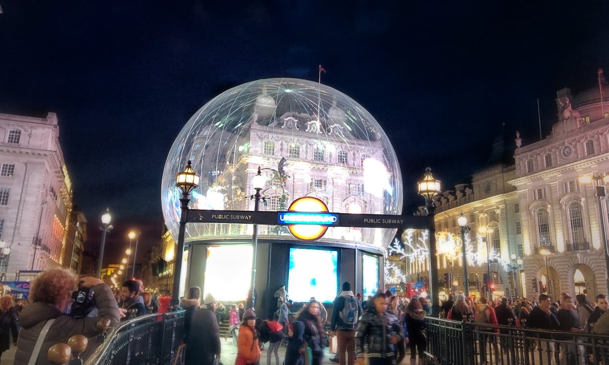 La boca de metro de Piccadilly Circus de noche, cubierta por una gran bola de nieve transparente de decoración navideña, con el logotipo del Underground iluminado debajo y la gente pasando alrededor.