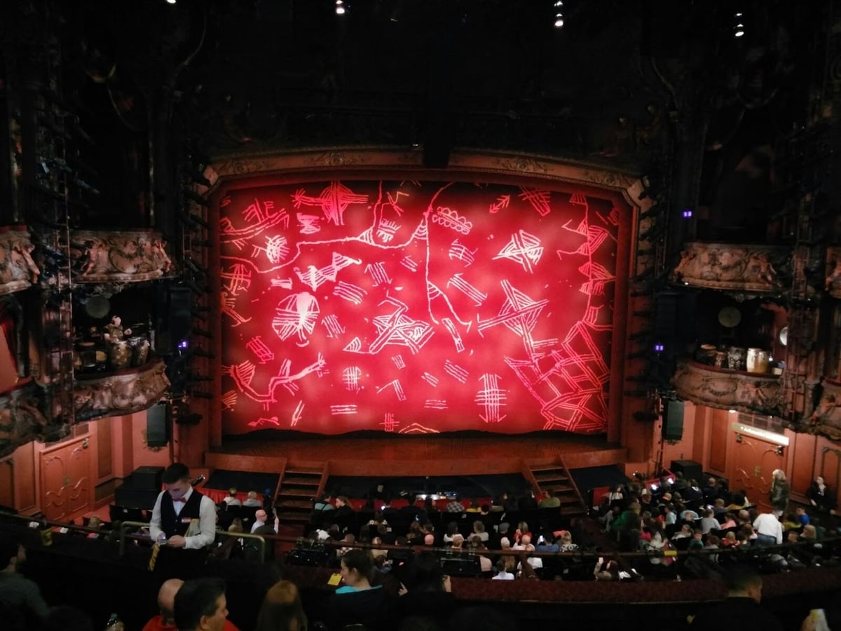 El telón rojo del Lyceum Theatre de Londres, decorado con trazos tribales en blanco y naranja, visto desde el anfiteatro, con los palcos ornamentados a los lados y el público ocupando sus butacas antes de la función.