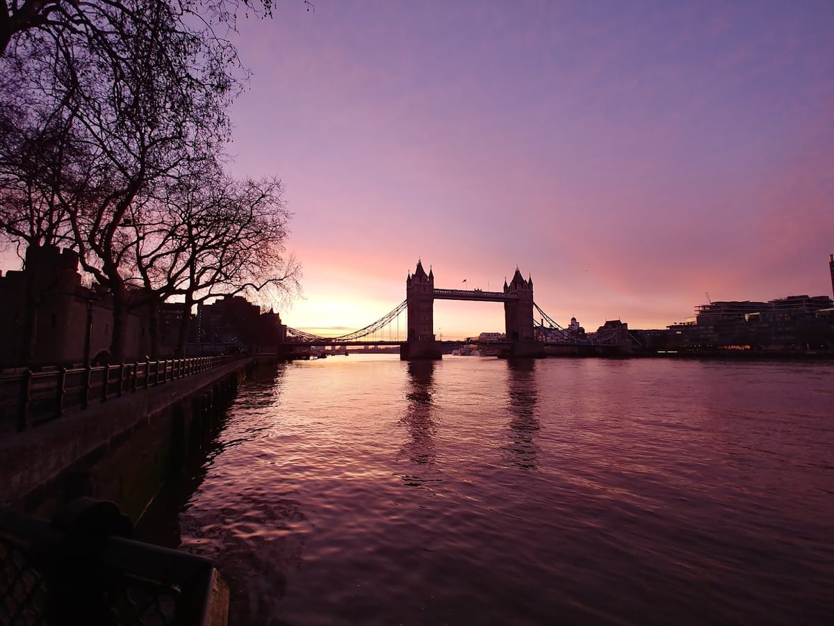 La Tower Bridge recortada en negro contra un amanecer rosa y malva sobre el Támesis, con la muralla de la Torre de Londres y unos árboles pelados en la orilla izquierda.