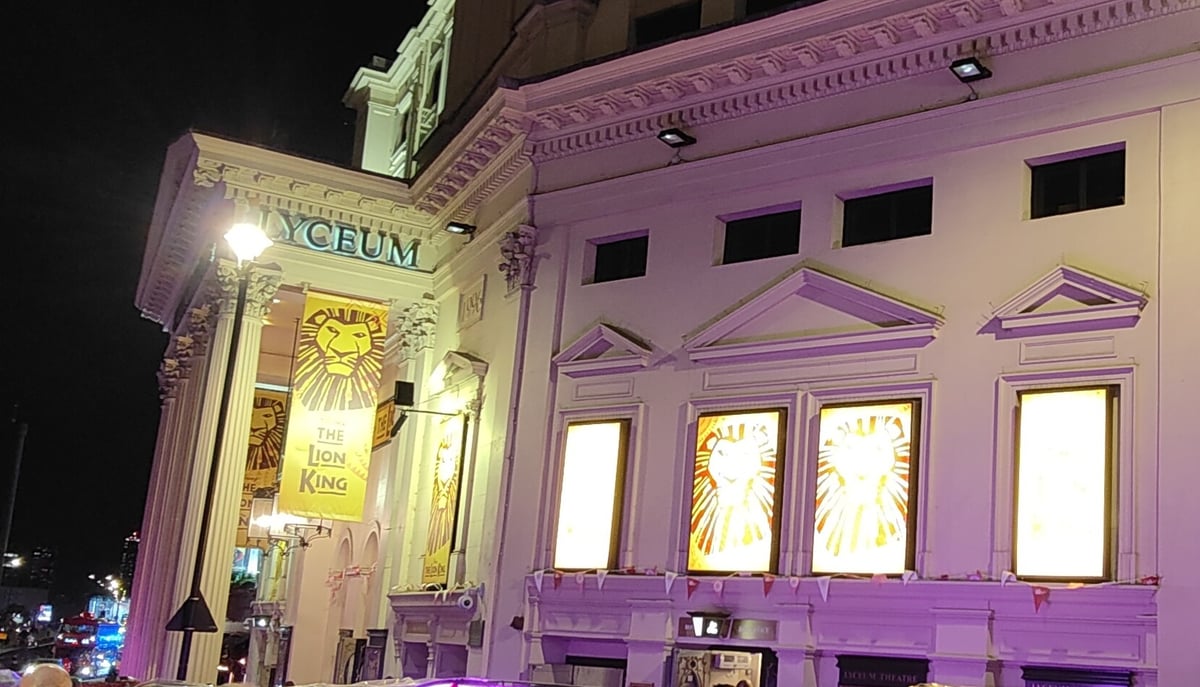 La fachada del Lyceum Theatre de Londres iluminada de noche, con el rótulo del teatro sobre las columnas, un estandarte amarillo de El Rey León colgado entre ellas y las vitrinas con la melena del león dibujada.