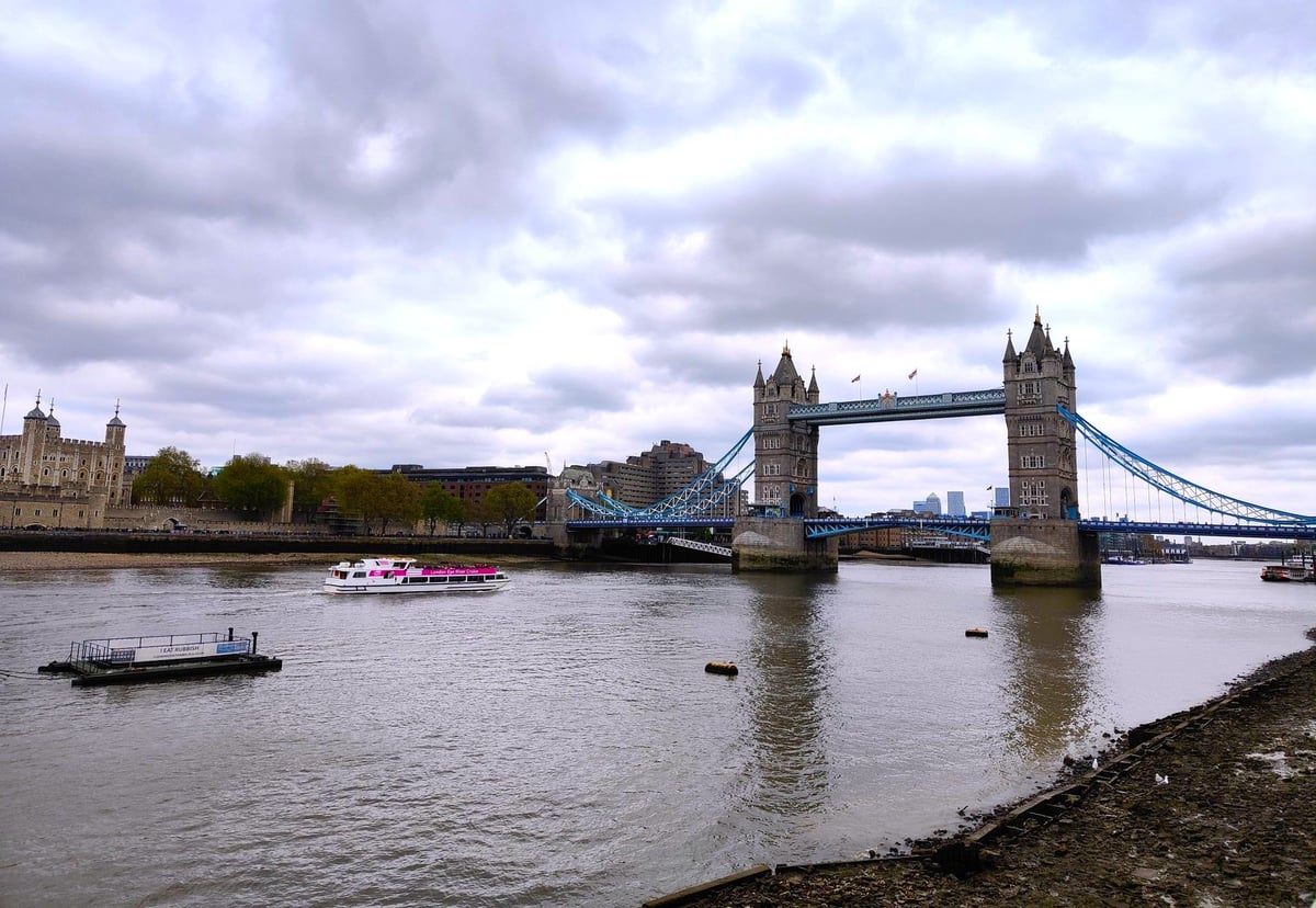 La Tower Bridge con sus tirantes azules bajo un cielo encapotado, vista desde la orilla del Támesis en marea baja, con la Torre de Londres a la izquierda y un barco turístico cruzando el río.