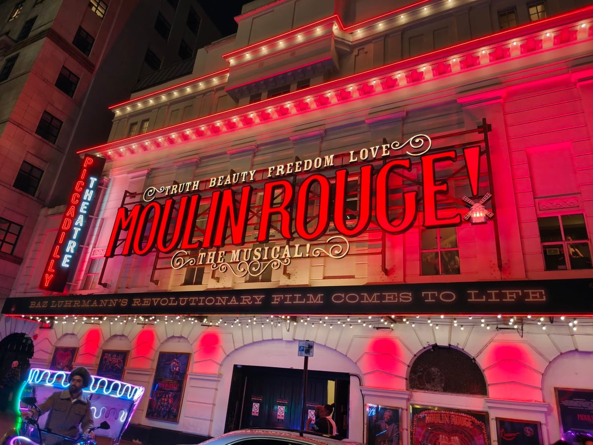 Fachada del Piccadilly Theatre de noche, iluminada en rojo, con el rótulo luminoso «MOULIN ROUGE! THE MUSICAL» bajo el lema «TRUTH BEAUTY FREEDOM LOVE», un molino de viento de neón a la derecha y el cartel vertical del teatro a la izquierda.