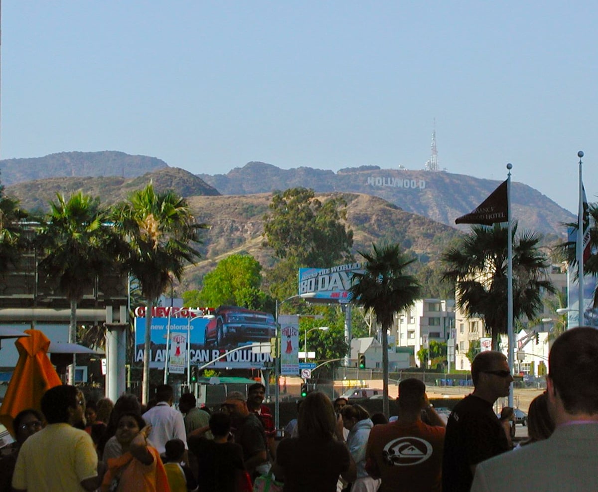 El rótulo de Hollywood en la ladera parda de las colinas, visto por encima de una multitud de espaldas, entre palmeras, vallas publicitarias y las banderas de un hotel.