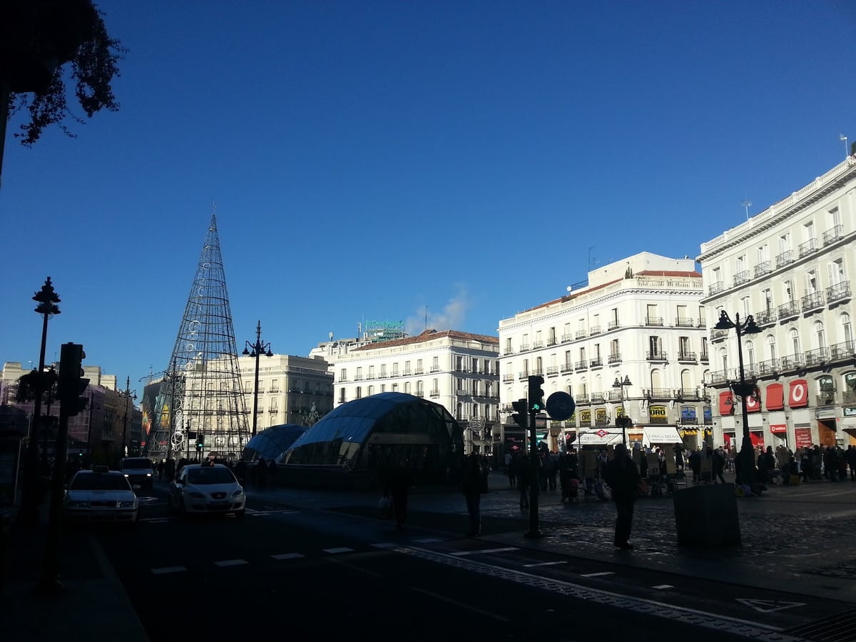 La Puerta del Sol de Madrid en Navidad, con el cono de luces del árbol a la izquierda, las cúpulas de vidrio de la estación de Cercanías en el centro y las fachadas blancas bajo un cielo azul intenso.