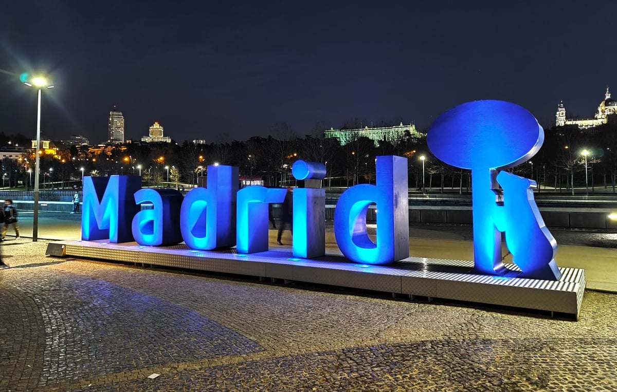 El rótulo escultórico de Madrid junto al río, con las letras iluminadas en azul y la última rematada por la silueta del oso y el madroño, y la ciudad encendida en la ladera del fondo.