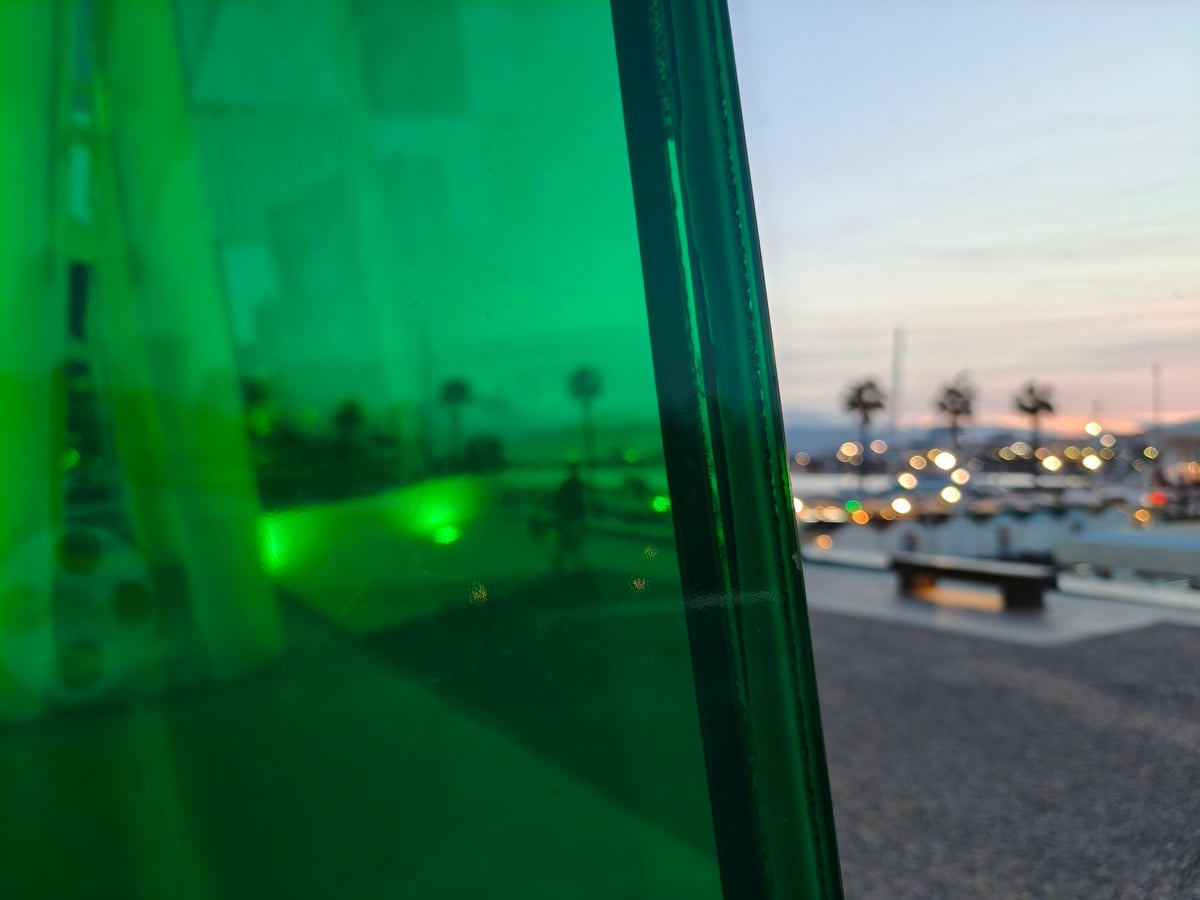Detalle del vidrio verde del Cubo del Centre Pompidou de Málaga en primer plano, con el puerto deportivo, las palmeras y las luces del muelle desenfocados al atardecer al otro lado del cristal.