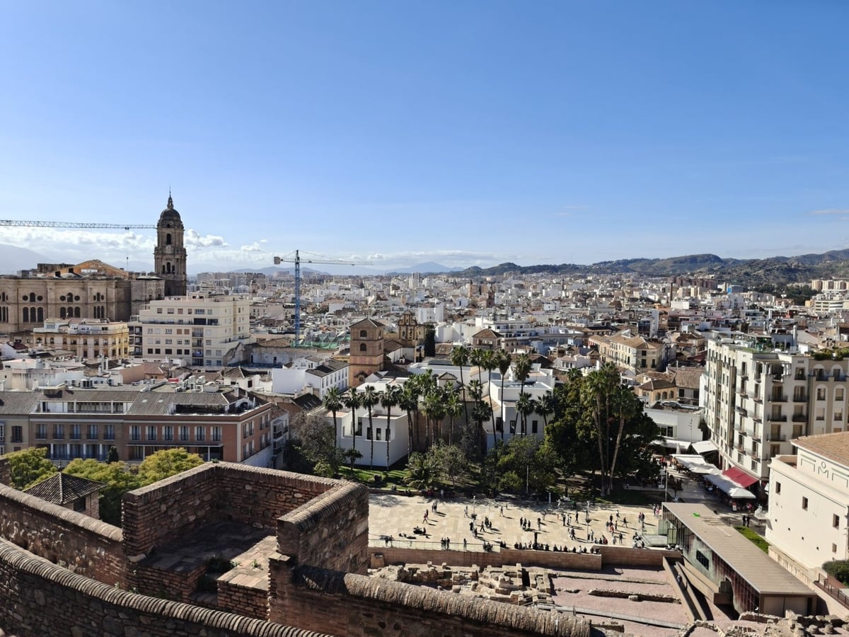 Málaga desde las murallas de la Alcazaba, con la torre de la catedral asomando a la izquierda, el caserío blanco extendiéndose hasta los montes y el teatro romano y sus palmeras justo debajo.