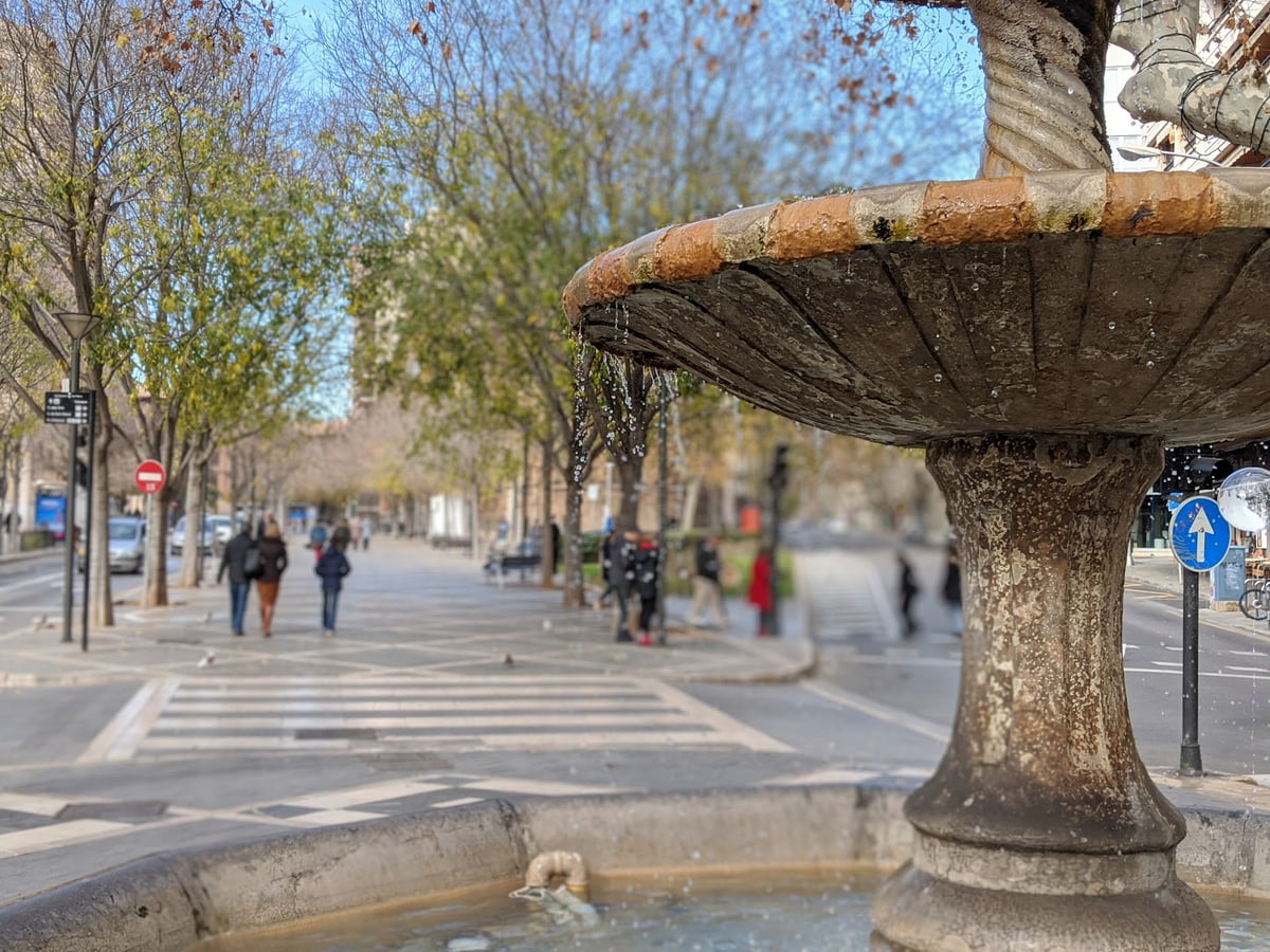 El pilón de piedra de una fuente en primer plano, con el agua rebosando por el borde, y detrás, desenfocado, un paseo arbolado con gente caminando en un día soleado de invierno.