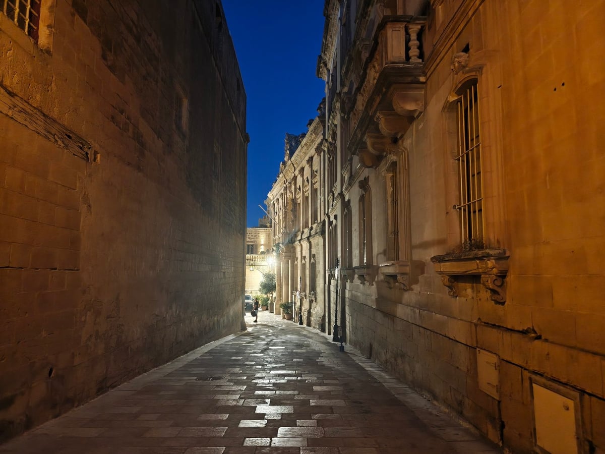 Una calle estrecha de Mdina vacía de noche, con los muros de piedra caliza iluminados por farolas.