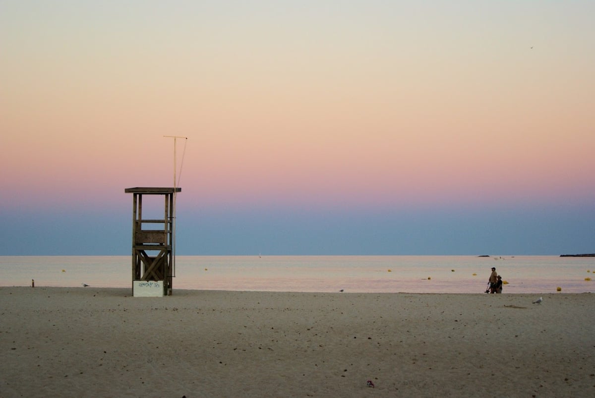 Una torre de vigilancia de madera sola en una playa de arena clara al anochecer, con el mar en calma y el cielo pasando del rosa al malva sobre el horizonte.